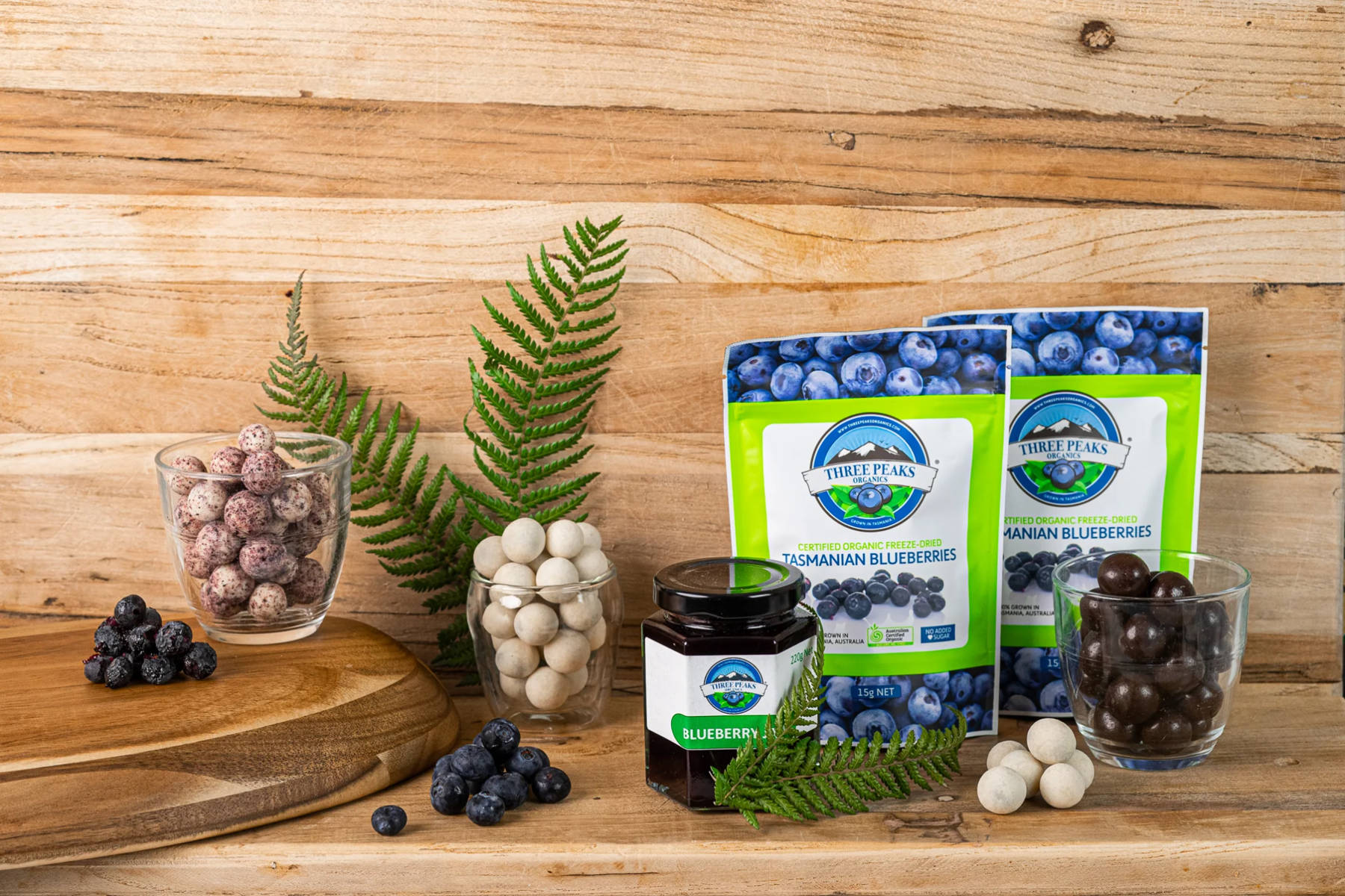 Various Three Peaks Organics products displayed on a wooden shelf and in glasses, with some loose blueberries and fern leaves. Photo: Three Peaks Organics.